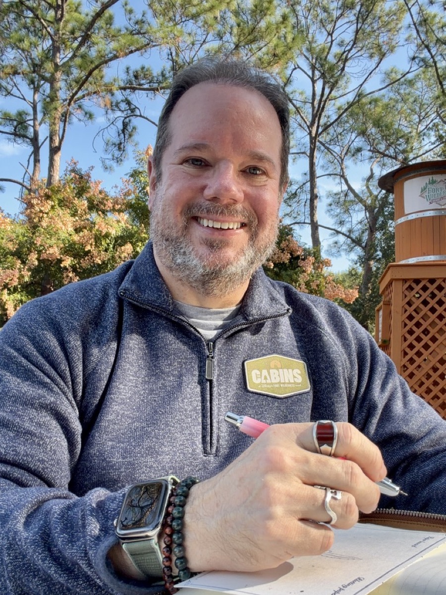 George smiling outdoors at Disney's Fort Wilderness Resort while writing in a notebook with a fountain pen.