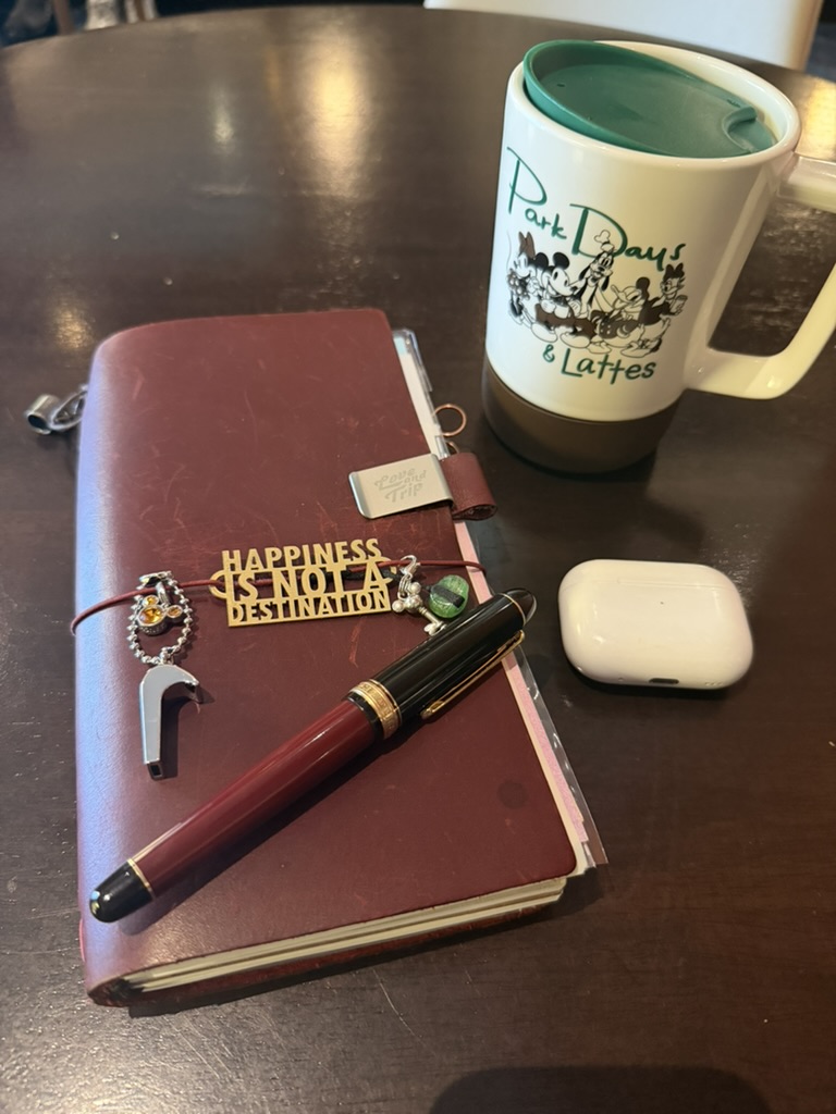 A Traveler's Notebook with charms, a Platinum 3776 Century Coffee Jelly fountain pen, earbuds, and a Parks Days and Lattes coffee mug on a table.